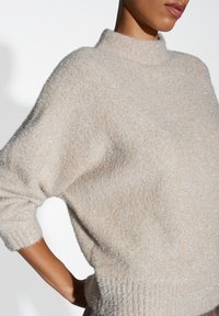 Pull beige en maille lâche avec un col montant, doté d'une texture douce et duveteuse ainsi que d'un léger scintillement. Détails de poignets et d'ourlet côtelés inclus.