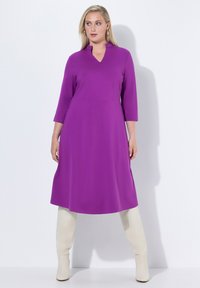 Robe violette avec un décolleté en V et des manches trois-quarts, fabriquée en tissu lisse, associée à des bottes blanches montantes. Lignes épurées et silhouette simple.