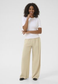 Haut blanc à manches courtes, pantalon beige léger à jambes larges avec poches avant, porté avec des chaussures noires. Une femme souriante pose avec une main sur son menton.