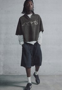 Olive green oversized short-sleeve shirt with graphic print, layered over a white long-sleeve shirt; paired with dark shorts and black sneakers.