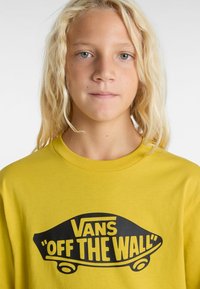Gul bomulls-t-shirt med en svart "VANS 'OFF THE WALL'" logografik på framsidan. Klassisk rundhalsdesign. Mjuk textur.