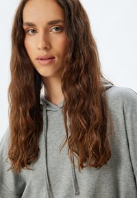 Sudadera gris con un corte relajado, que presenta una capucha con cordón, una textura suave y un sutil patrón jaspeado. Se puede ver el cabello ondulado de longitud media.