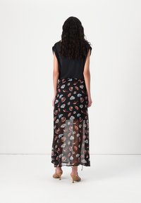 Femme aux longs cheveux bouclés portant un haut noir sans manches et une jupe noire transparente avec des motifs de coquillages rouges et blancs, debout sur des talons beige.