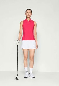 Rosa pikétröja med krage, ihop med en vit plisserad kjol. Svart golfklubba hålls i ena handen, bär vita sportstrumpor och skor.