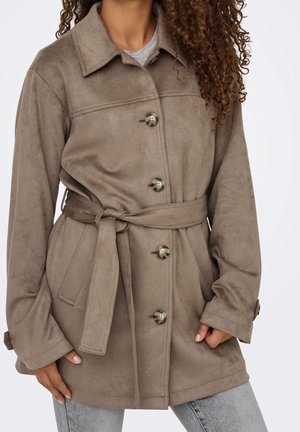 Personne aux cheveux bouclés portant un manteau taupe ceinturé avec quatre boutons, debout les mains près des poches sur un jean gris clair.