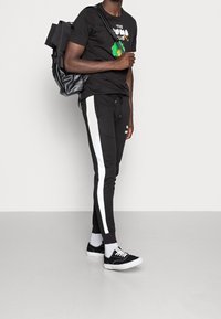 Camiseta negra con estampado gráfico, pantalones de chándal negros con rayas laterales blancas, mochila negra y zapatillas negras con suelas blancas.