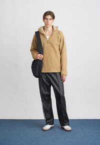 Beige fleece pulóver fél cipzárral és kapucnival, fekete műbőr nadrággal, amelyen fehér oldalsávok találhatók, és fehér sportcipőkkel.