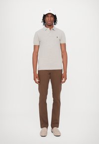Polo grigio a maniche corte, con piccolo logo ricamato verde, pantaloni marroni e scarpe slip-on beige chiaro, completato da un cappellino bianco.