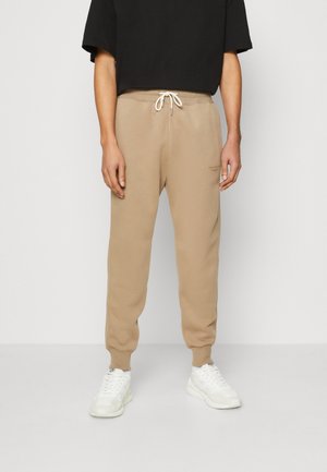 Personne portant un pantalon de jogging beige avec un cordon blanc, un t-shirt noir à manches courtes et des baskets blanches, debout devant un fond uni.