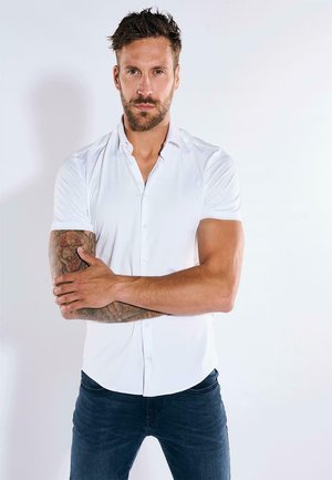 Weißes kurzärmeliges Hemd mit Knopfverschluss, tailliertem Schnitt, glatter Textur und abgerundetem Saum. Modell trägt dunkle Jeans und hat sichtbare Armtattoos.