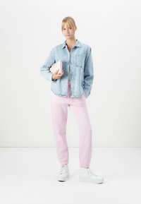 Jeune femme portant une veste en denim clair, un pantalon de survêtement rose, des baskets blanches, tenant une pochette rose pâle, debout contre un fond blanc uni.