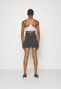 Sujeción deportiva gris con tirantes finos, combinada con pantalones cortos deportivos negros que tienen un dobladillo ligeramente acampanado y un pequeño logo blanco en la cinturilla.