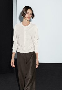 Una donna in un cardigan beige chiaro e pantaloni scuri cammina con sicurezza, incorniciata da sfondi in contrasto tra chiaro e scuro.