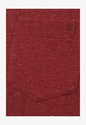 Tissu rouge avec une poche texturée et un petit logo d'arbre brodé dans le coin supérieur droit de la poche.