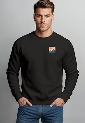 Mann mit kurzen Haaren, der einen schwarzen Pullover mit kleinem Grafikmotiv von Bergen und Sonne trägt, Hände in den Taschen der blauen Jeans, neutraler grauer Hintergrund.