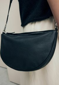 Sac à épaule en cuir noir avec une forme arrondie, un extérieur texturé, une fermeture zippée en haut et une bandoulière réglable.