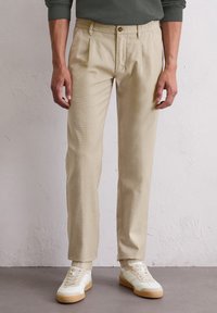 Homme portant un pantalon beige plissé et des baskets blanches, debout contre un mur texturé blanc.
