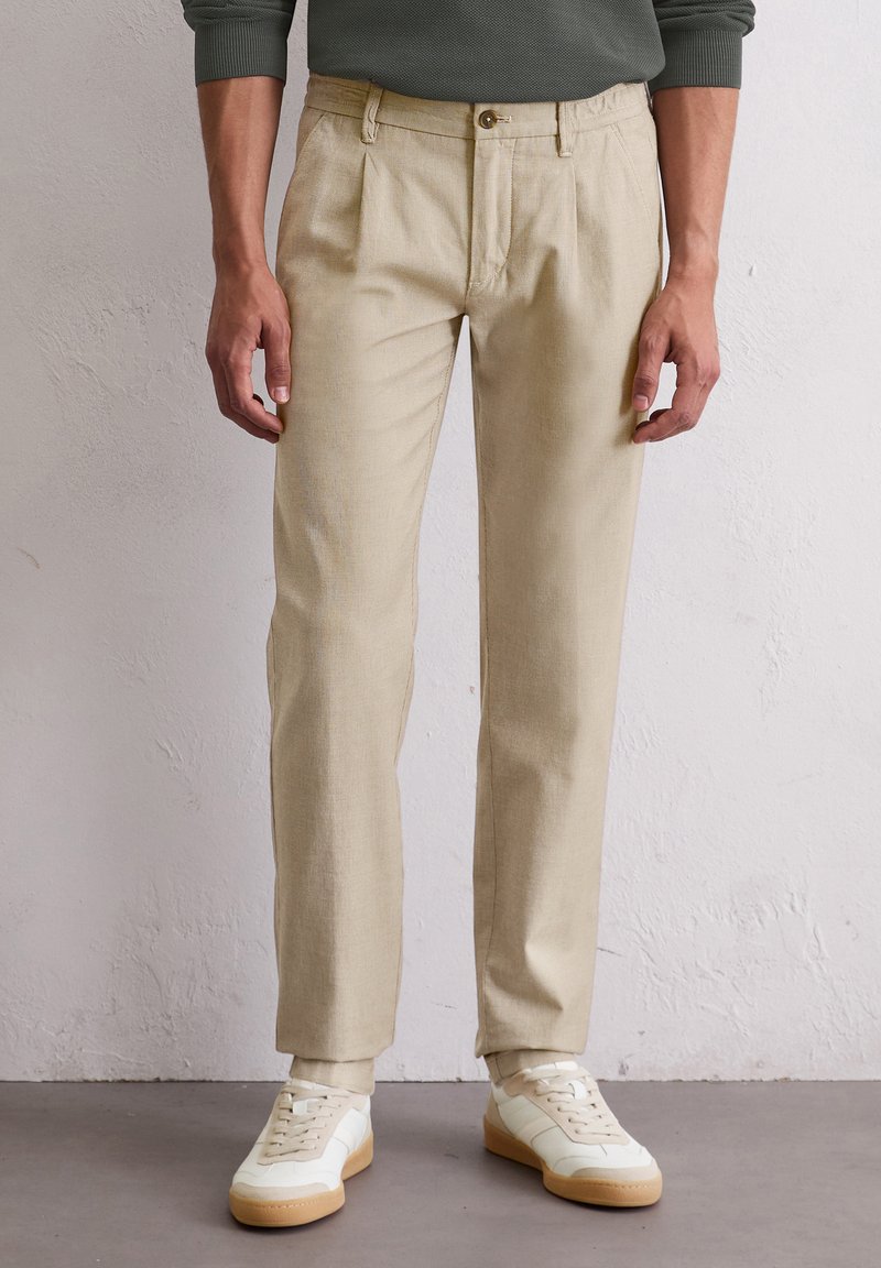 Homme portant un pantalon beige plissé et des baskets blanches, debout contre un mur texturé blanc.
