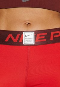 Červené legíny Nike Pro mají široký hnědý pas s výrazným červeným textem "NIKE PRO" a bílou štítkem zobrazujícím podrobnosti o produktu.