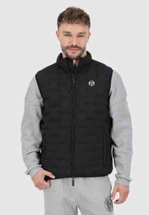 Schwarze gepolsterte Weste mit hohem Kragen, Reißverschluss und Logodetail auf der Brust; getragen über einem grauen Langarmshirt.