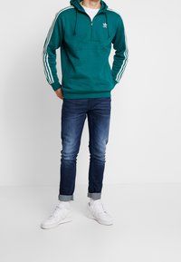 Tyrkysová mikina na zip s bílými třemi pruhy a logem, v kombinaci s modrými džíny a bílými teniskami. Uvolněný střih s žebrovanými manžetami.