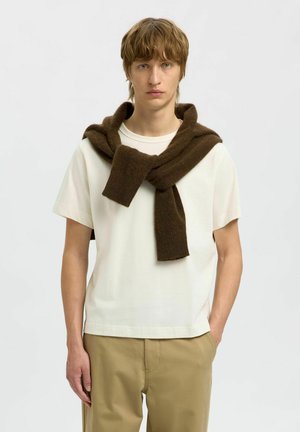 Brauner Wollschal, der über einem weißen T-Shirt drapiert ist, kombiniert mit hellbeigen Hosen. Der Schal ist dick, hat eine weiche Textur und ein asymmetrisches Design.