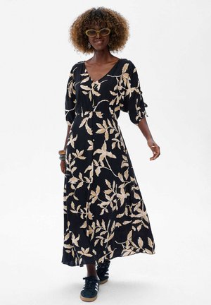 Vrouw met krullend haar in een zwart-beige bloemenmaxi-jurk, groene cat-eye zonnebril en zwarte sneakers die vooruit loopt.