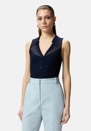 Donna con top blu navy a coste senza maniche con bottoni e pantaloni a vita alta color azzurro chiaro, in piedi davanti a uno sfondo chiaro.