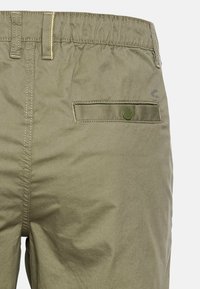 Pantalons kaki avec une taille élastique, une poche arrière plaquée avec fermeture à bouton et des détails de couture visibles en fil contrasté.