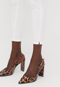Pointed toe high heels with a glossy leopard print finish, featuring a slingback design and block heel, worn with white ankle length pants.