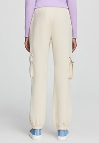 Kremowe spodnie cargo z kieszeniami bocznymi, elastycznym pasem i mankietami. Miękka tkanina o gładkiej fakturze. Noszone z niebieskimi sneakersami.