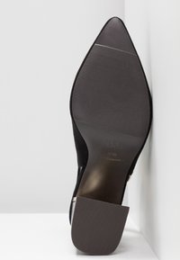 Semelle d'une chaussure noire à bout pointu avec semelle antidérapante texturée et talon carré, taille 37/38, posée contre un coin de fond blanc.