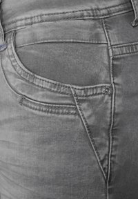 Jeans de mezclilla gris con un primer plano de los detalles del bolsillo trasero, que presentan una textura suave, costuras sutiles y un acento de remache metálico.