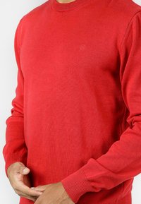 Czerwony sweter dziewiarski z okrągłym dekoltem, ściągaczami przy mankietach i gładką fakturą. Zawiera subtelną naszywkę z logo na piersi.