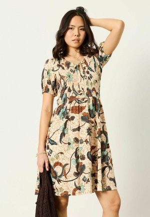 Young woman in floral patterned dress with puff sleeves, holding a brown textured fabric in one hand, posing with other hand on head.