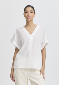 Blouse blanche, légère, texturée avec un col en V, des manches courtes et une coupe ample, associée à un pantalon de couleur claire, à coupe décontractée.