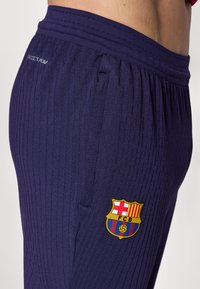 Lilla sportbukser med ribbet tekstur, med et gult og rødt FC Barcelona-logo på siden og en tætsiddende talje.