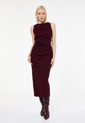 Bordeaux mouwloze jurk met een aansluitende silhouette, textuurstof, knielange snit en subtiele zijrimpeling, gecombineerd met bruine enkellaarsjes.
