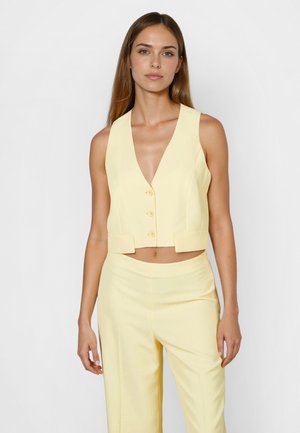 Woman wearing a sleeveless pale yellow cropped vest with three buttons and matching high-waisted pants against a plain background.