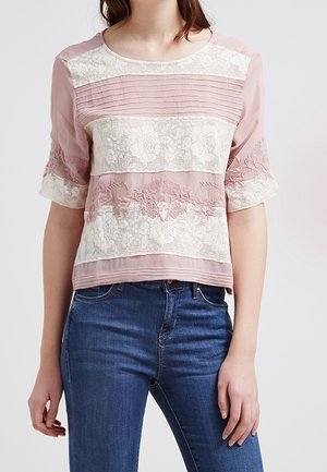 Kvinde iført lyserød bluse med hvide blondepaneler og blå jeans, stående mod en ensfarvet lys baggrund.