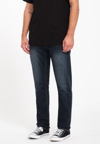 Jeans de mezclilla de un azul oscuro con corte recto, combinados con zapatillas negras. La tela tiene una textura suave y los jeans presentan detalles de costura mínimos.