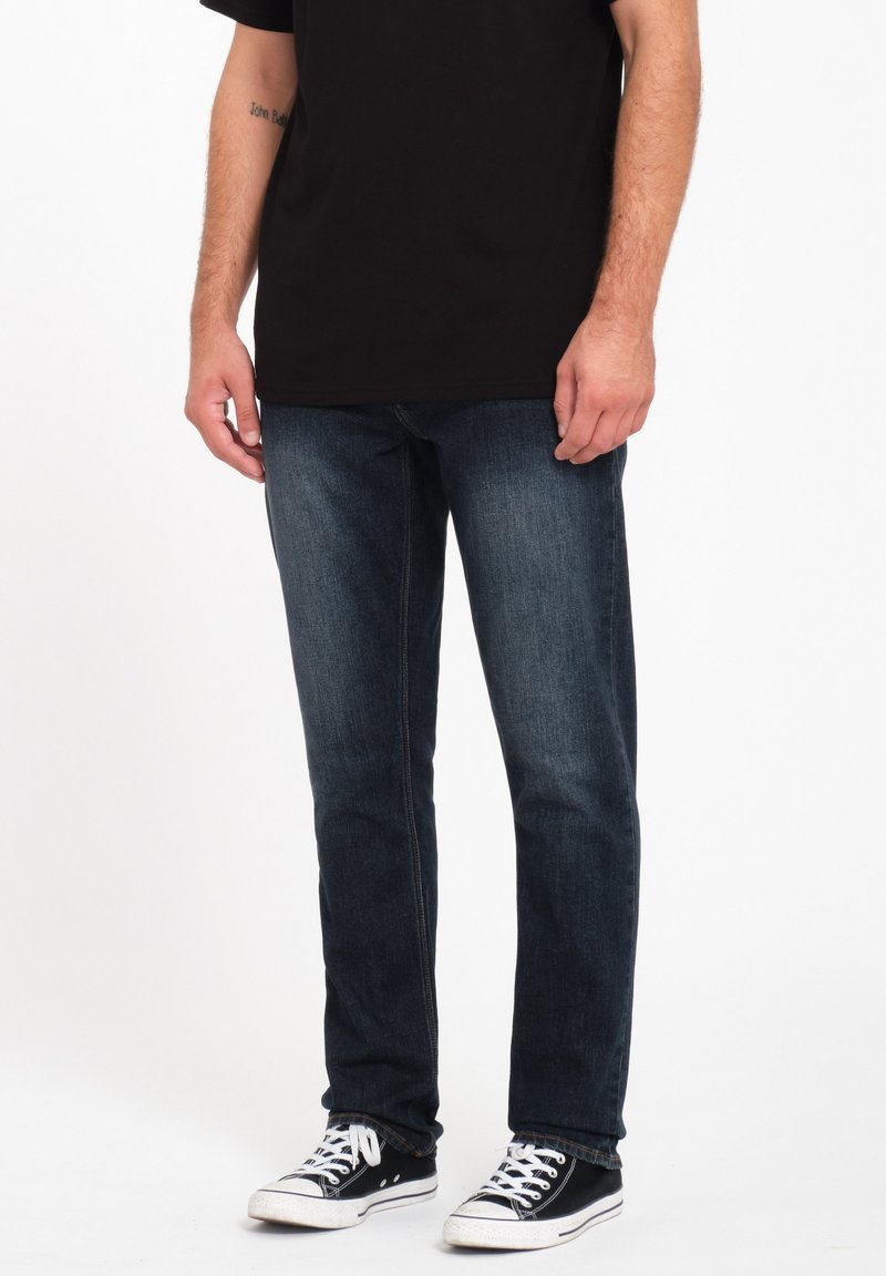 Jeans de mezclilla de un azul oscuro con corte recto, combinados con zapatillas negras. La tela tiene una textura suave y los jeans presentan detalles de costura mínimos.
