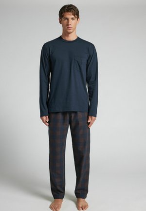 Junger Mann steht barfuß, trägt ein marineblaues Langarmshirt mit Brusttasche und dunkle karierte Pyjamahosen vor einem schlichten Hintergrund.