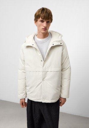 Veste blanche rembourrée avec une capuche zippée, des boutons-pression et une couture horizontale. Portée sur un haut gris clair. Pantalon noir texturé.