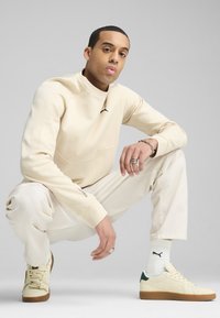 Sudadera de color beige claro con cuello alto y logo negro, combinada con pantalones beige claros y zapatillas crema con detalles verdes.