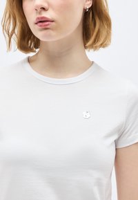 Νέα γυναίκα με κοντά κόκκινα μαλλιά, φορά απλό λευκό t-shirt με μικρό ασημένιο καρφίτσα "8" στο στήθος και ασημένιο κρίκο σκουλαρίκι.