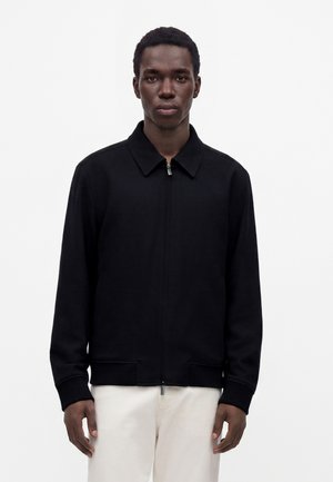 Jeune homme portant une veste noire zippée avec col et un pantalon blanc, debout devant un fond clair uni.
