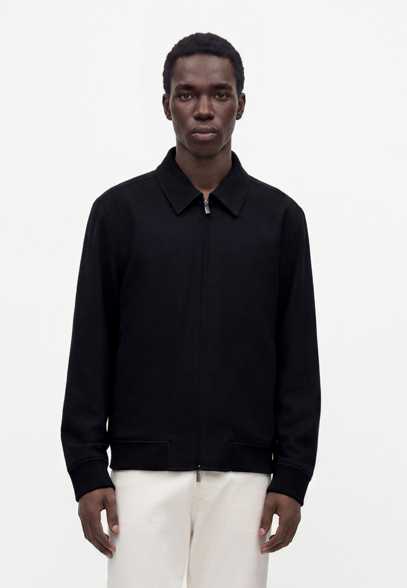 Jeune homme portant une veste noire zippée avec col et un pantalon blanc, debout devant un fond clair uni.