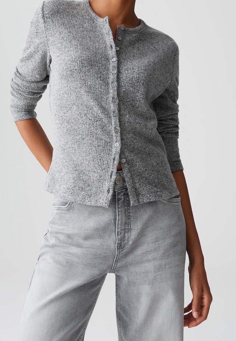 Femme portant un cardigan en maille gris boutonné à manches longues et un jean taille haute gris clair sur un fond uni.