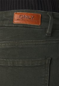 Gros plan d’un jean en denim vert foncé avec un patch en cuir marron portant l'inscription « ONLY blue denim since 1995 » au-dessus de la poche arrière.